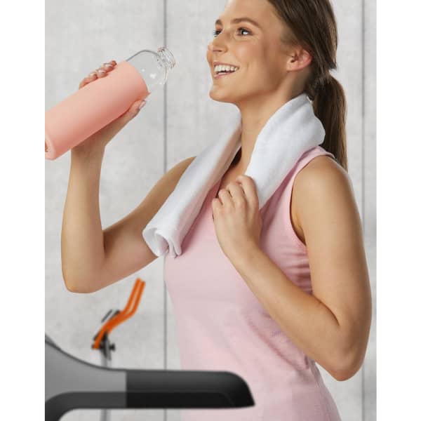 A woman in a pink top holds a clear drink bottle while wearing a white towel around her neck, set in a gym environment.