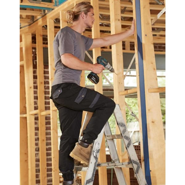 A pair of black work pants on a model using a power drill while standing on a ladder amidst wooden framing.