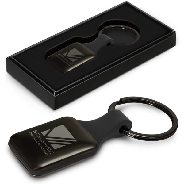 A square black key ring with a shiny surface, presented in a gift box. Features a logo on the key ring.