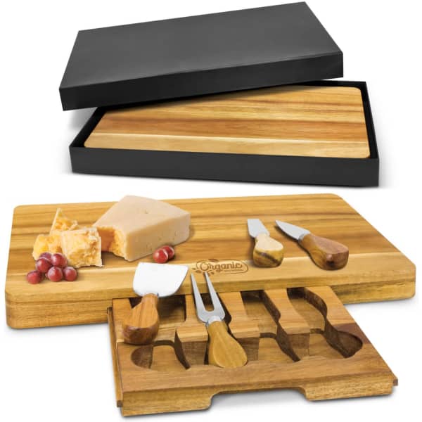 A wooden cheese board with three cheese knives and a black storage box. The board is light brown.
