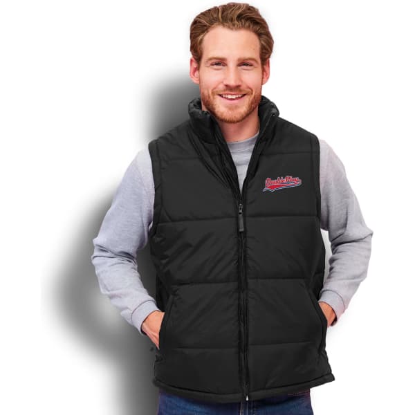 A black unisex puffer vest with a zip-up front and a logo. The model is smiling and wearing it over a grey top.