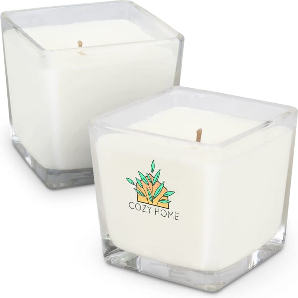 A square, white scented candle in a clear glass container, featuring a logo design. Two candles are shown.