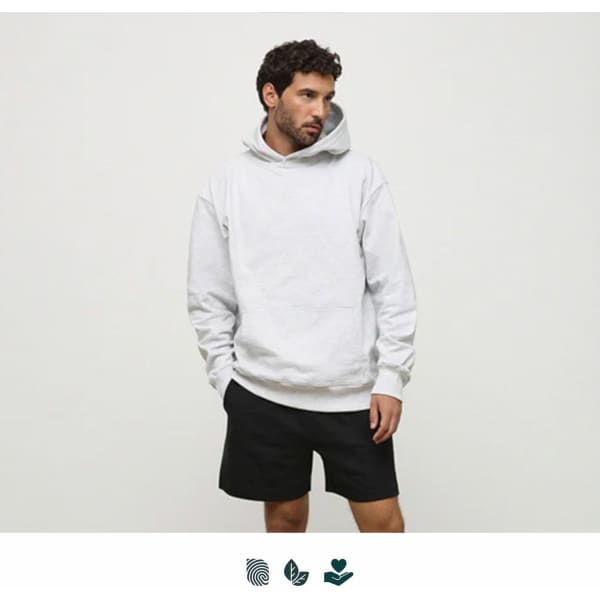 A man wearing a light grey hoodie and black shorts stands against a plain background. The hoodie has a logo.