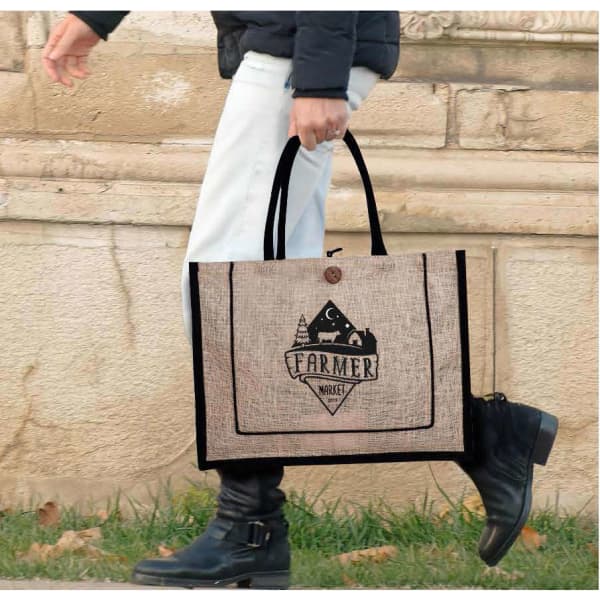A person carrying a beige jute bag with black accents and a logo on the front, outdoors on a path.