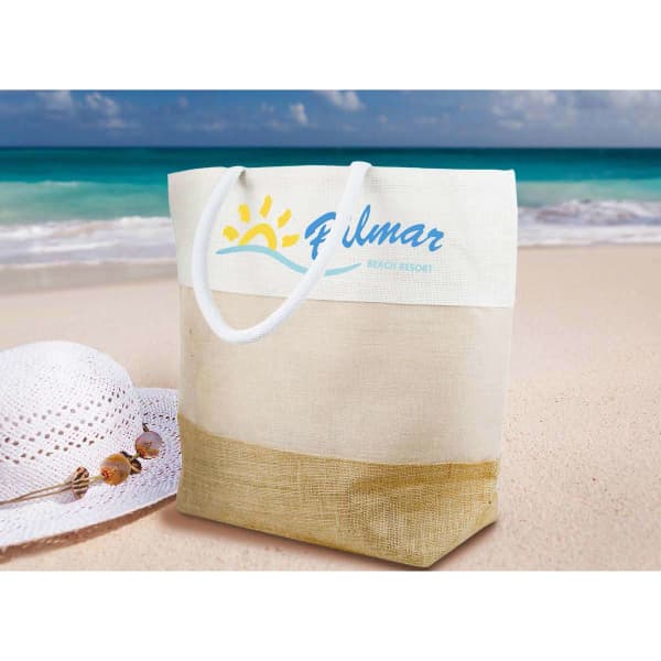 A cream and tan beach bag with a logo, set against a sandy beach and ocean backdrop.