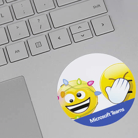A round sticker featuring emojis and a logo, placed on a silver laptop keyboard. The sticker has bright colours.