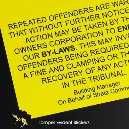 A close-up of a bright yellow tamperproof vinyl sticker with a logo, displaying text about violations and penalties.
