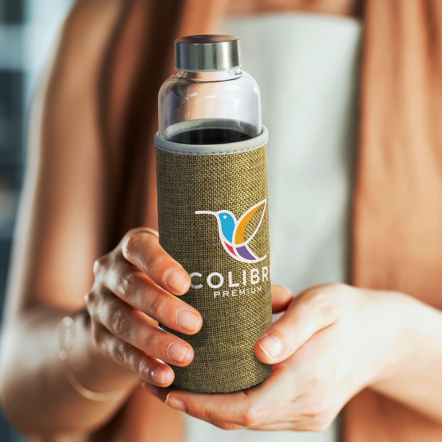 A drink bottle with a clear glass body and a green natural sleeve, held by a person with an outstretched hand.