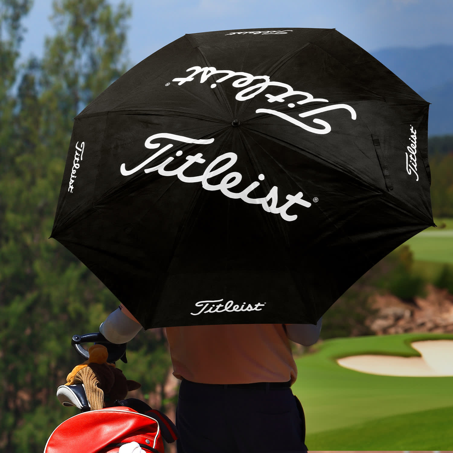 A black double canopy umbrella with a logo, held by a person near a golf course.