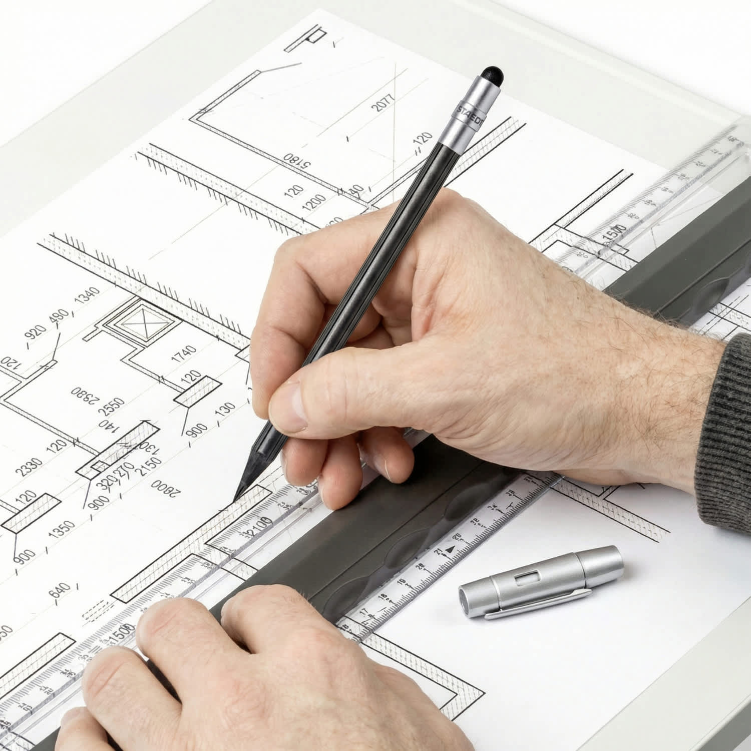 A hand holding a grey pencil sketches on a blueprint with a ruler and a silver mechanical pencil nearby.