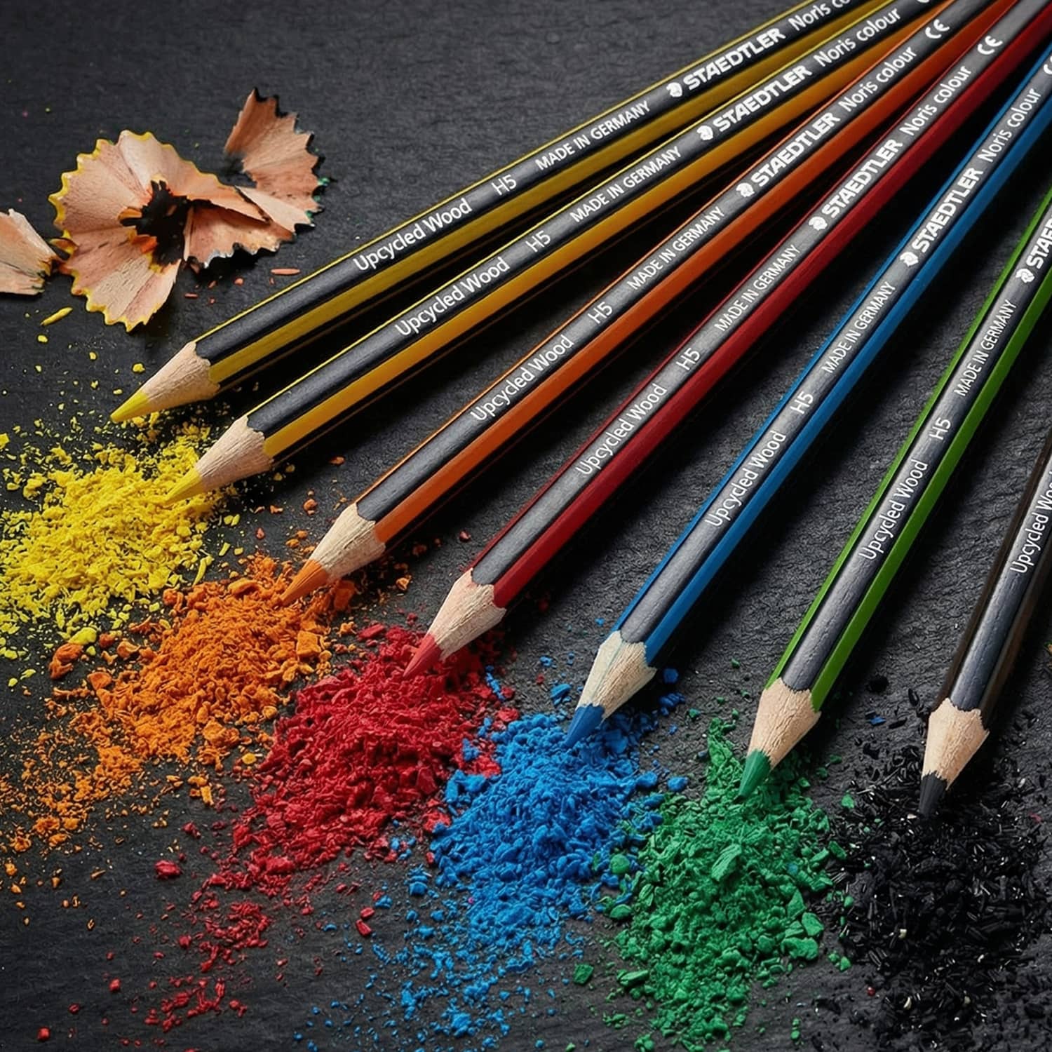 A set of six coloured pencils in yellow, orange, red, blue, black, and green, with shavings and colour powder scattered around.