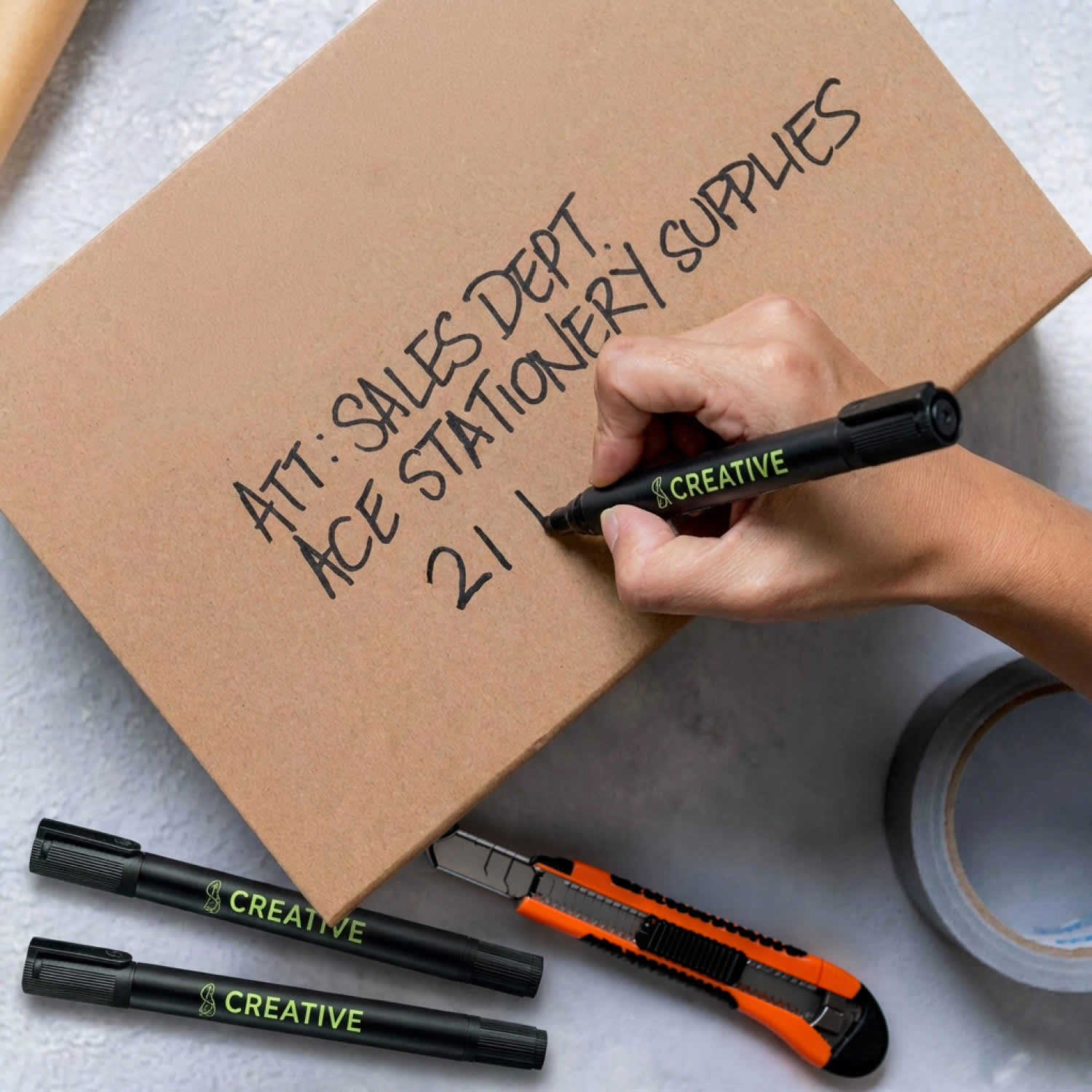 A hand writes on a cardboard box using a black Staedtler Lumocolor permanent marker with a chisel tip.