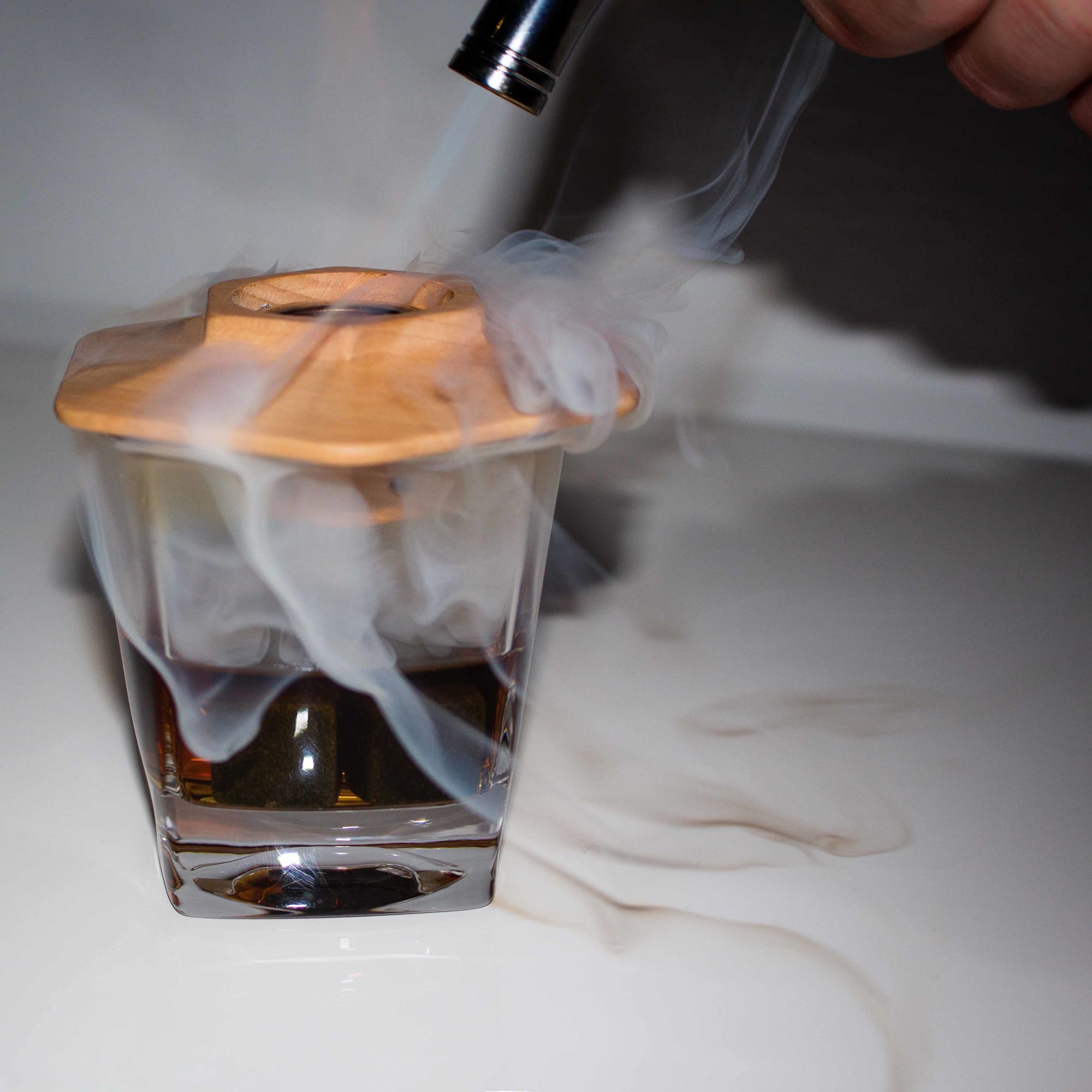 A whiskey glass filled with amber liquid, topped with a wooden lid, emitting wisps of smoke from a smoking device.