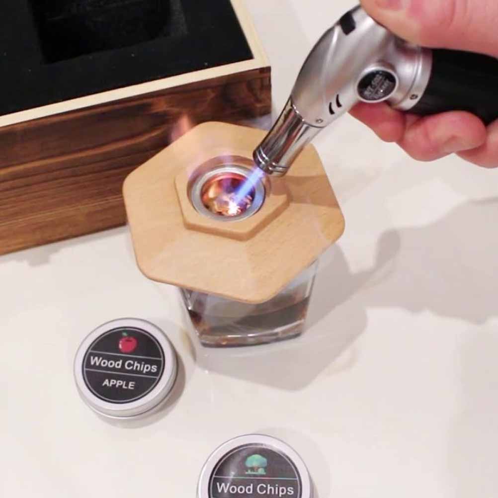 A wooden smoker lid with a torch igniting wood chips in a glass jar, alongside two containers labelled ‘Apple Wood Chips’.