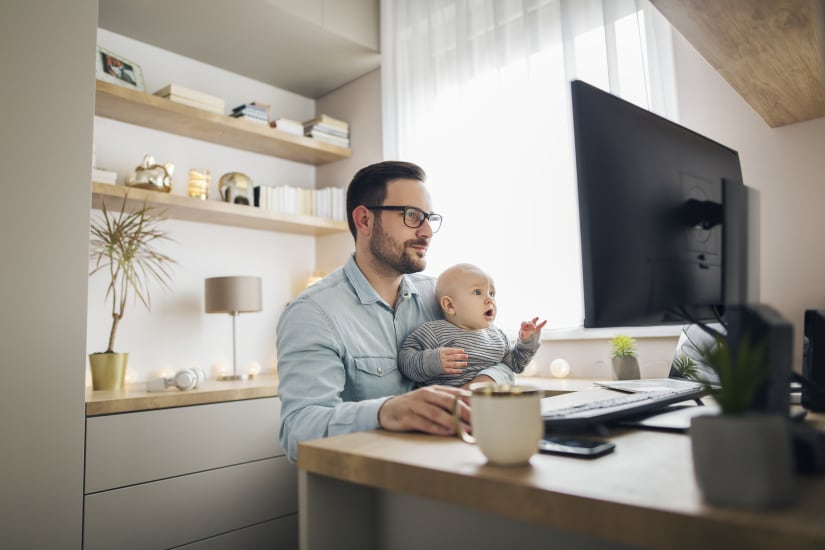 A man is working at his home office while he holds his baby in his laptop.