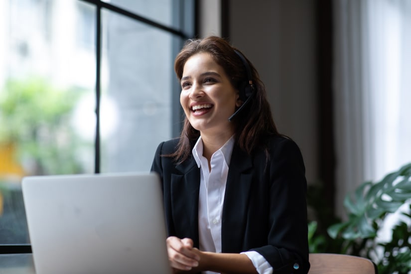 A woman wearing a headset and business outfit is smiling while looking above her laptop.