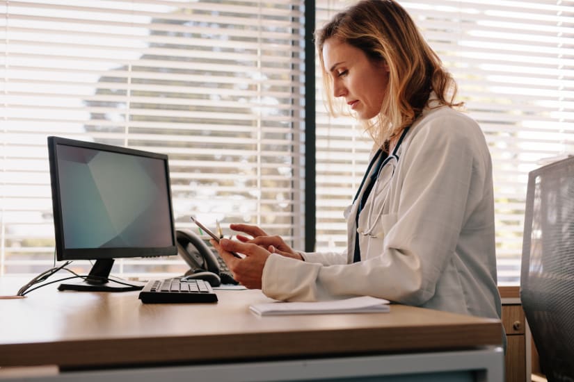 A doctor checks something on her phone while seated next to her computer.