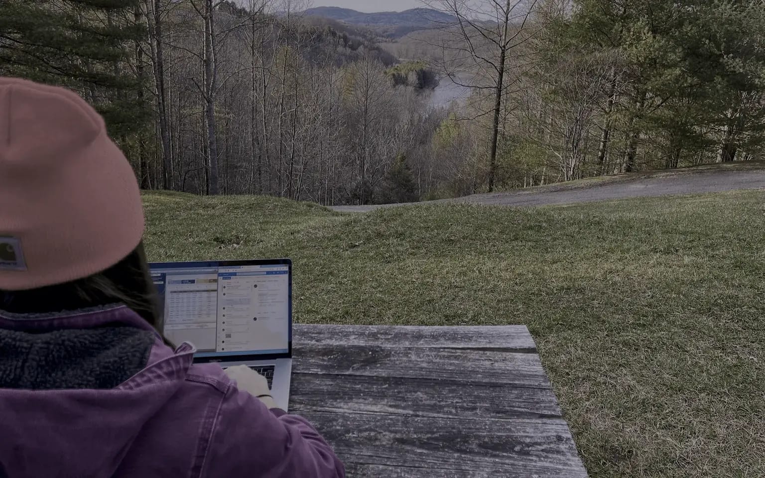 A woman uses Intuit QuickBooks on her laptop at a picnic table in a wooded mountain area.