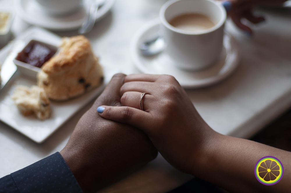 close-up photograph of hand holding, near a table with scones and tea, highlighting the woman's engagement ring