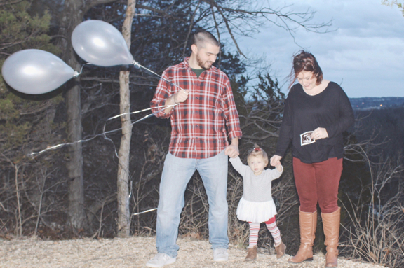 Rose standing between Mike and Becky. Becky is holding an ultrasound over her stomach, Mike holding two silver balloons.
