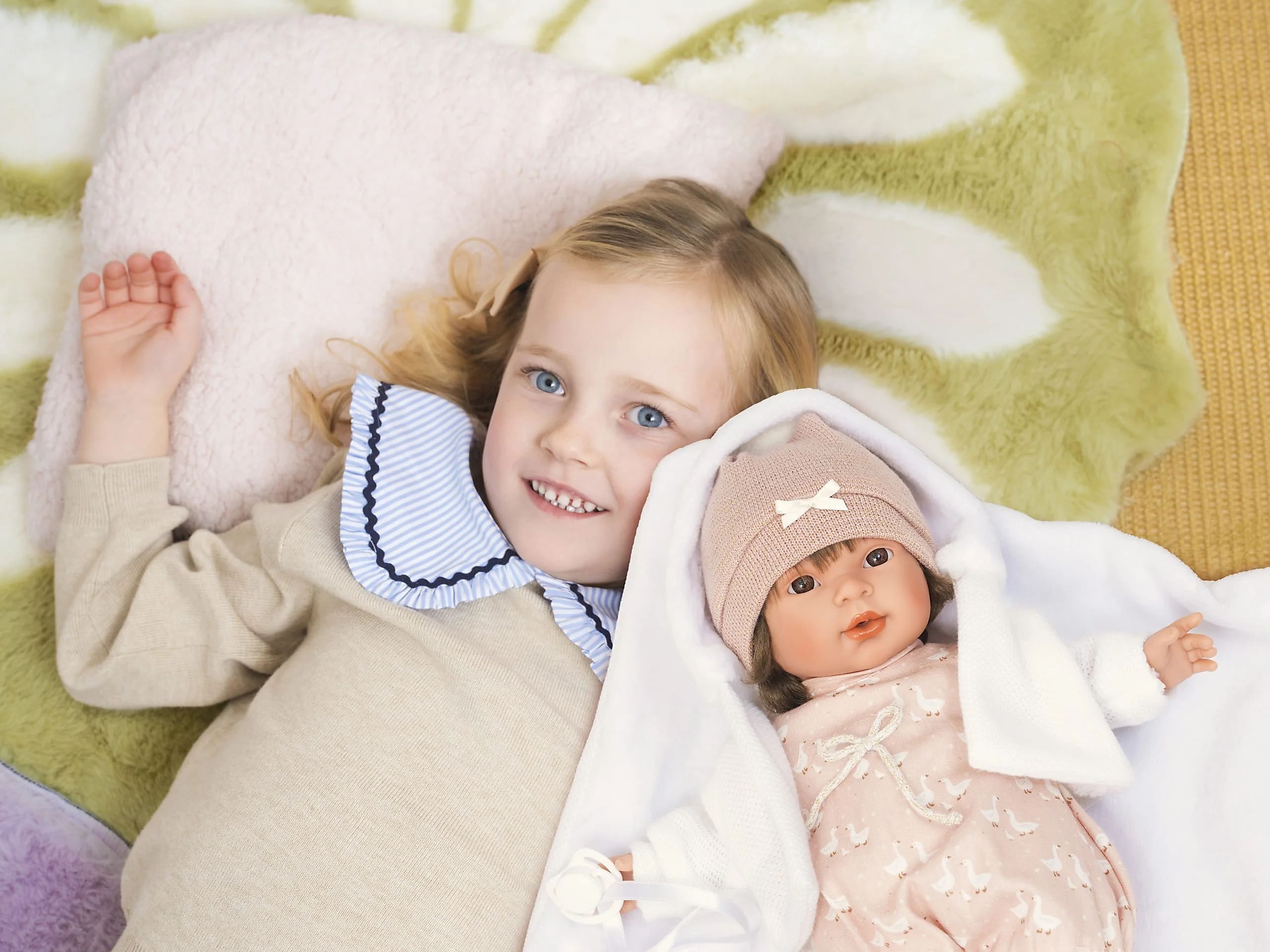 A child lying on a rug beside an Arias baby doll wrapped in a white blanket