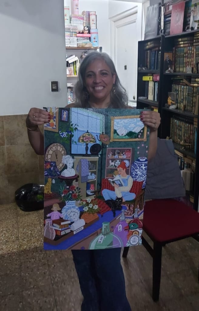 A woman holds up a vibrant illustration of a cozy, book-filled room with a person reading and a black cat.