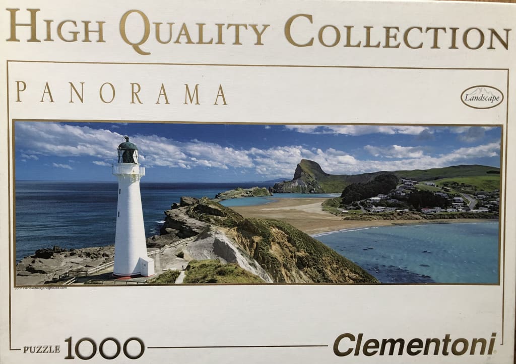 A panoramic coastal landscape featuring a white lighthouse overlooking a beach and rocky cliffs under a blue sky.