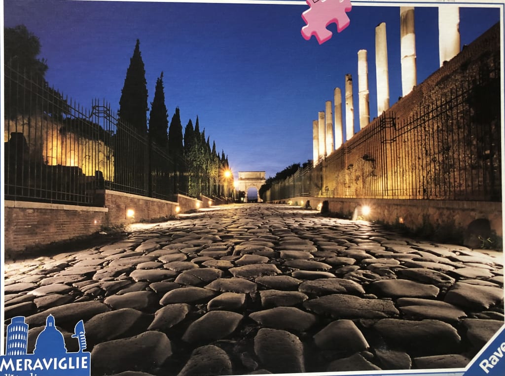 Night view of a Roman cobblestone street lined with ancient columns and cypress trees leading to an archway.
