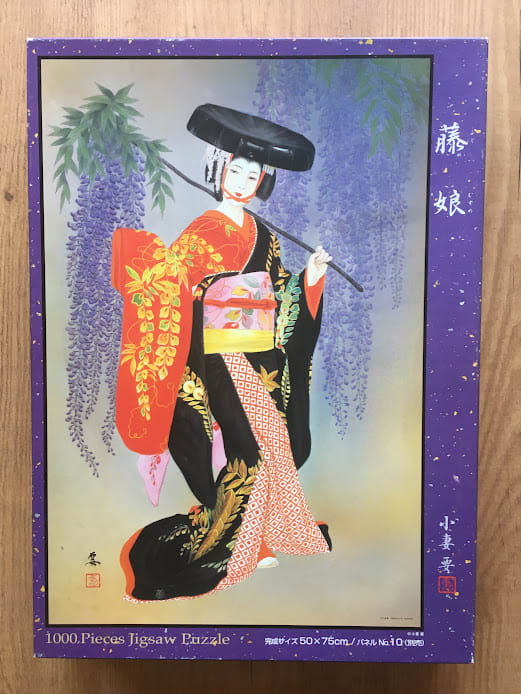 A traditional Japanese woman in a vibrant kimono stands beneath purple wisteria blossoms.