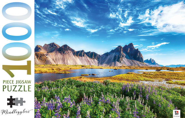 Vibrant landscape of a field of purple lupine flowers in front of a calm lake and jagged mountains under a blue sky.