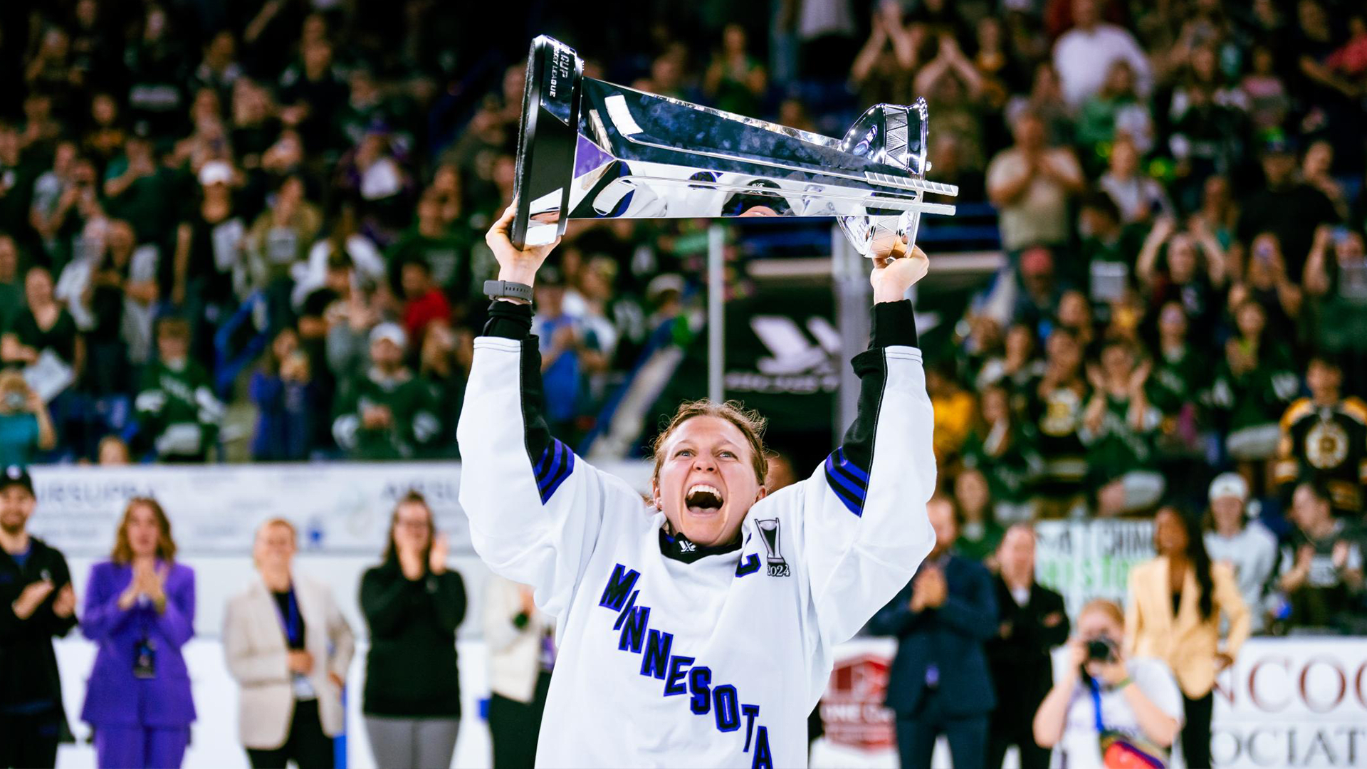 Minnesota's Captain Kendall Coyne Schofield, who is a blonde woman wearing a white and purple hockey uniform, cheering as she lifts the Walter Cup in front of the crowd. People cheering and clapping are visible in the background.