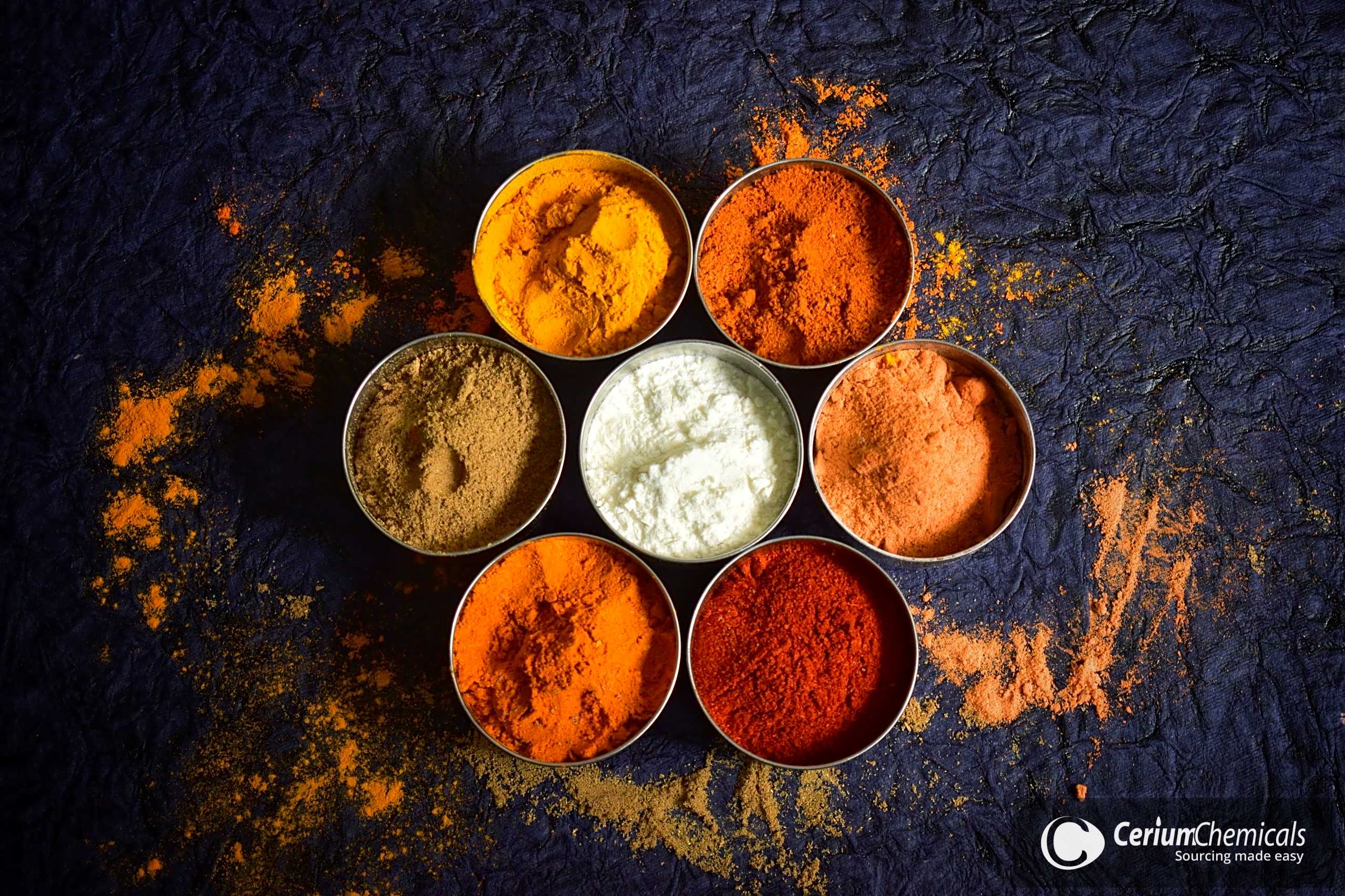Six metal tins of coloured spice and milk powders arranged on dark slate.