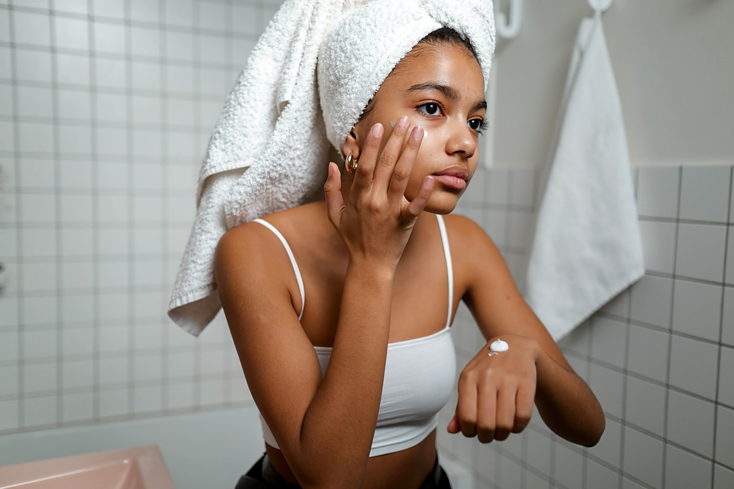 A woman smoothing cream onto her cheek in a white-tiled bathroom.