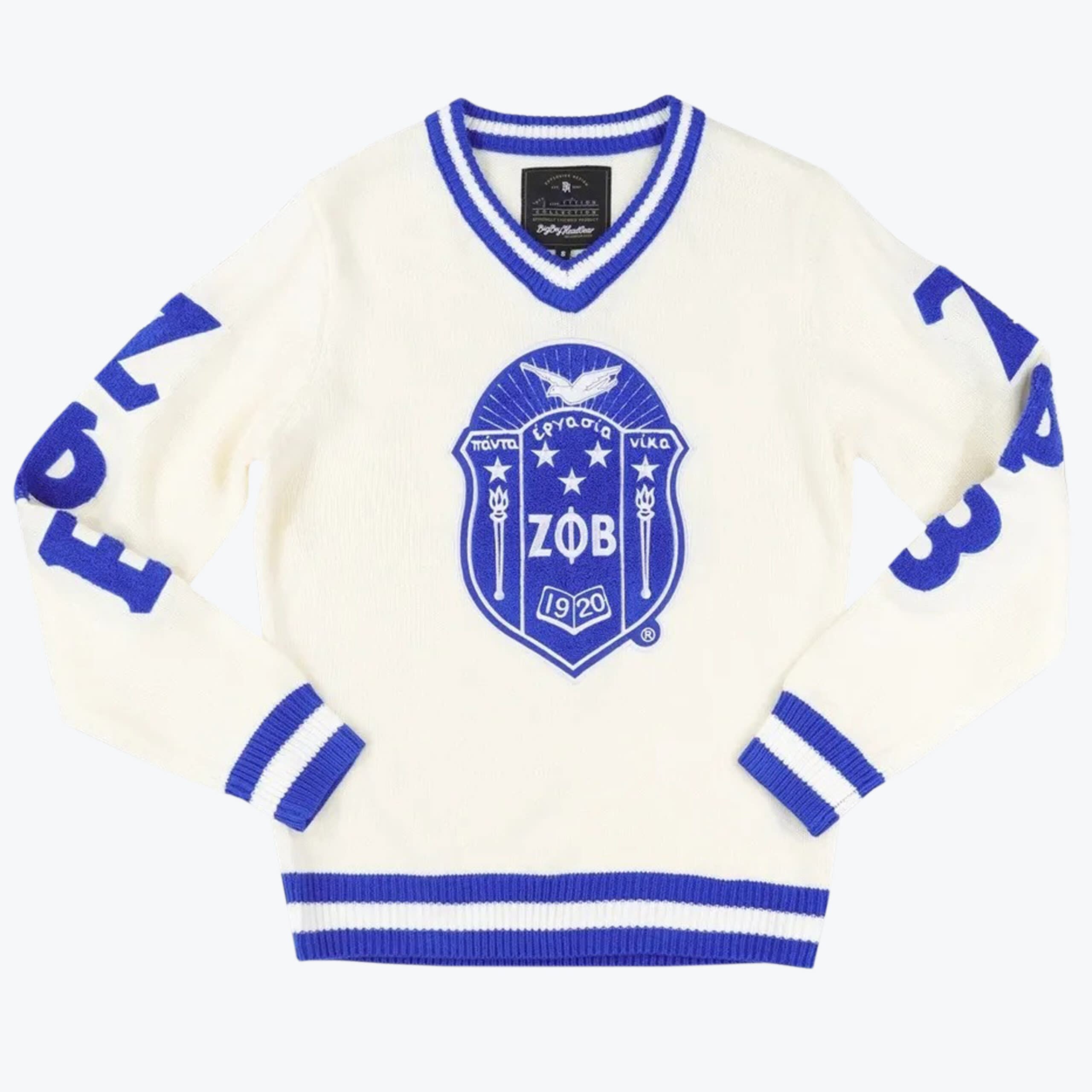 Zeta Phi Beta V-Neck Knit Sweater An off-white knit V-neck sweater with royal blue striped trim, featuring a large Zeta Phi Beta sorority shield emblem in the center and blue lettering on the sleeves.