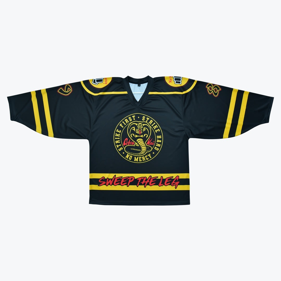 Cobra Kai Sweep the Leg Hockey Jersey A black hockey jersey featuring the yellow Cobra Kai logo in the center and Sweep the Leg in red script across the bottom.