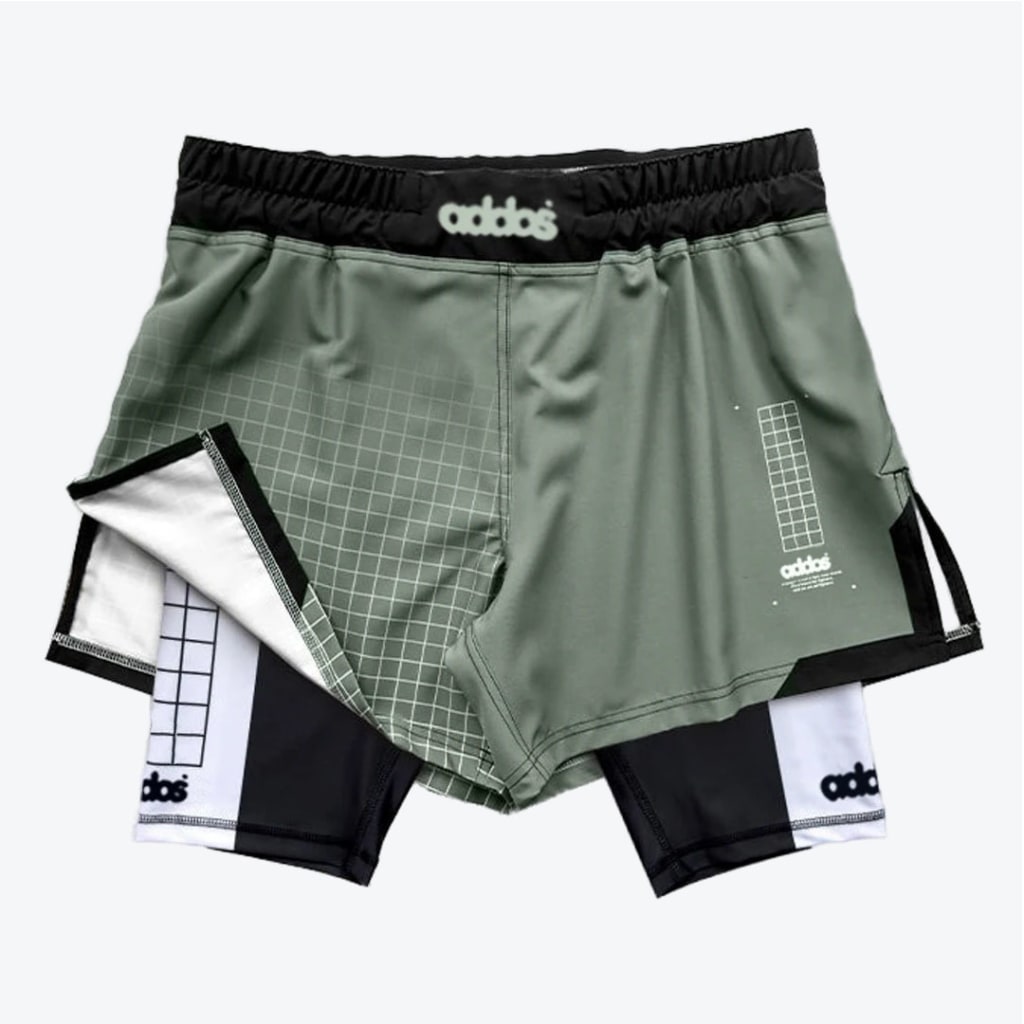 A pair of sage green two-in-one athletic shorts with a black and white inner compression layer, featuring grid patterns and stylized branding.
