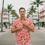 A man in a red floral Hawaiian shirt adjusting his collar while standing on a boardwalk with palm trees and Art Deco buildings in the background.