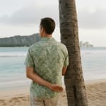 Rear view of a man in a green tropical print shirt leaning against a palm tree on a beach, looking towards the ocean and distant mountains.