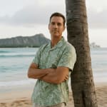 A man in a green Hawaiian shirt leans against a palm tree on a sandy beach with Diamond Head in the background.