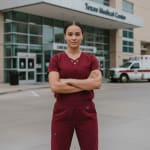 A female healthcare professional in maroon scrubs stands with crossed arms in front of the Texas Medical Center building, with an ambulance in the background.