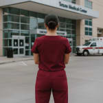 A rear view of a healthcare worker in maroon scrubs standing in front of the Texas Medical Center with an ambulance in the background.