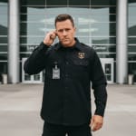 A security guard in a black uniform with a gold badge and earpiece stands in front of a modern building with glass doors.
