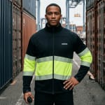 A man wearing a black and neon yellow high-visibility safety jacket stands between rows of shipping containers at a port, holding a walkie-talkie.