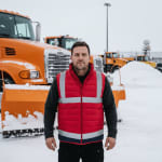 A male snowplow driver wearing a red reflective safety vest stands in front of an orange snowplow truck in a snowy depot.