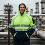 A man wearing a neon yellow and navy blue high-visibility safety hoodie with reflective stripes stands in front of an industrial refinery.