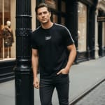 A young man in a black t-shirt and dark jeans leaning against a black decorative street pillar in an urban setting.