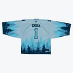 Back view of a light blue hockey jersey with CHIGA and the number 1 printed on it, featuring a navy blue pine tree forest silhouette pattern along the bottom and sleeves.