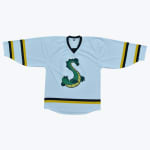 A flat lay of a light blue hockey jersey with a green sea serpent logo shaped like the letter S on the chest, detailed with black and gold stripes on the sleeves and hem.