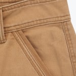 Close-up of the waistband, belt loop, and pocket of a pair of tan denim pants, showing the detailed twill texture and stitching.