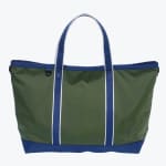 An olive green canvas tote bag with navy blue handles and base, featuring white stripes on the handles and a small D-ring on the front, isolated on a white background.